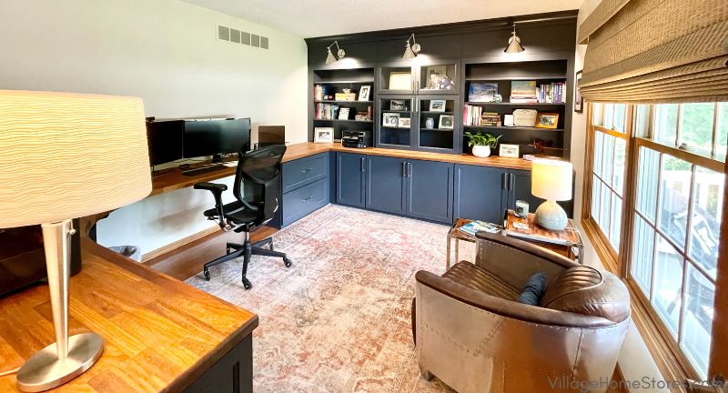 Wide shot of home office with U-shaped wood desktop and painted blue cabinetry with open bookshelves and glass door section.