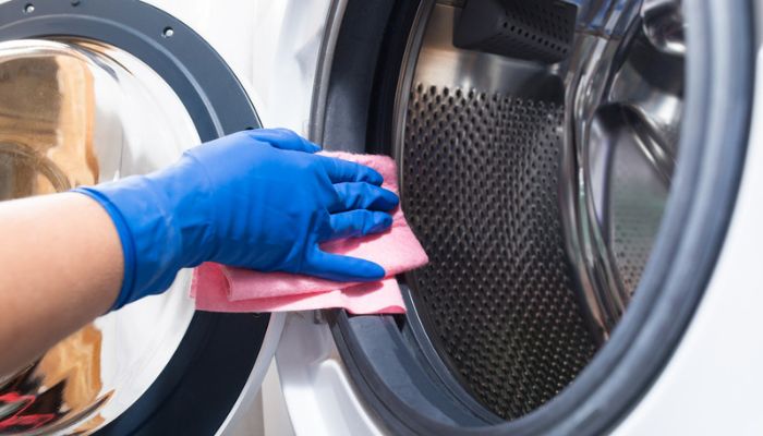 Gloved hand uses pink cloth to clean inside of an empty dryer