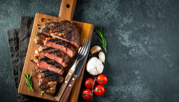 A steak is sliced on a wooden cutting board with utensils, herbs, garlic, and tomatoes