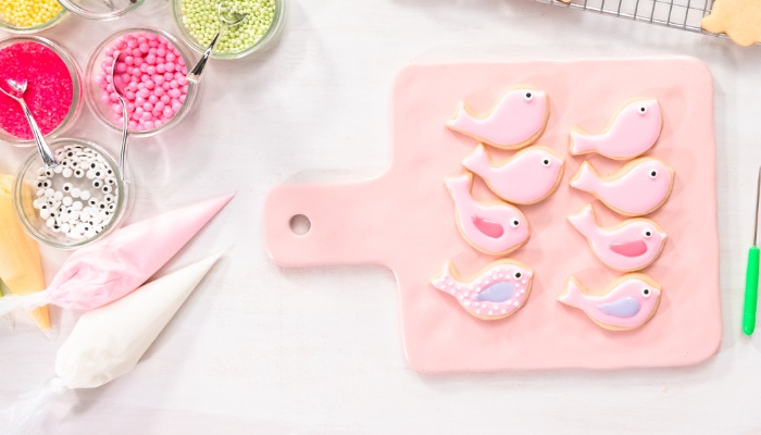 A workspace has bowls of sprinkles, bags of icing, and a pink cutting board with pink bird sugar cookies on it.