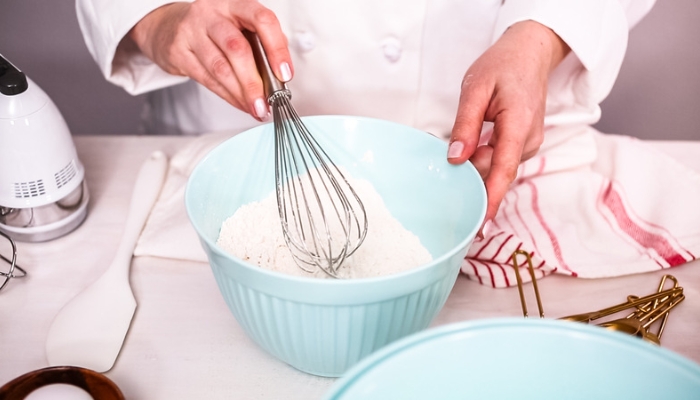 A chef mixes flour with a whisk in a blue ceramic bowl.