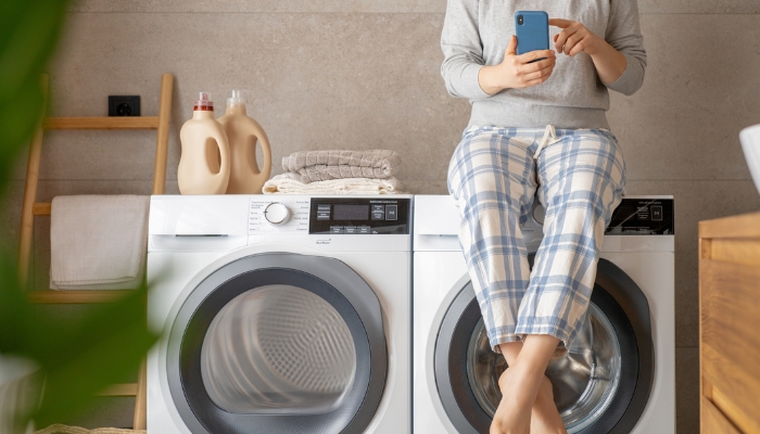 Woman sits on top of a washing maching with phone and next to detergent