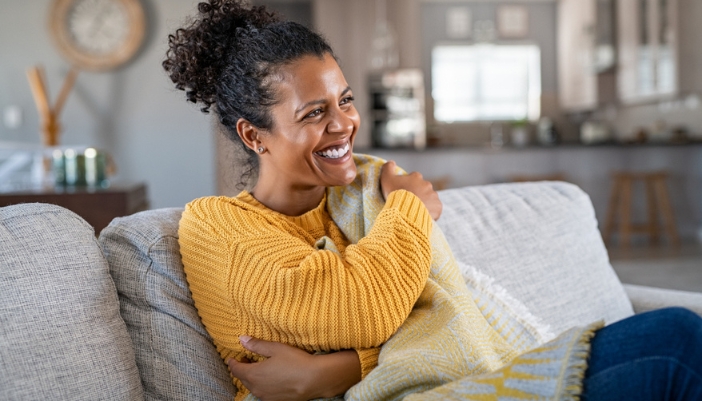 A woman sits on a gray couch and hugs a warm yellow blanket.