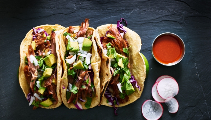 Three shredded tacos with toppings rest on a gray smooth surface with sauce.
