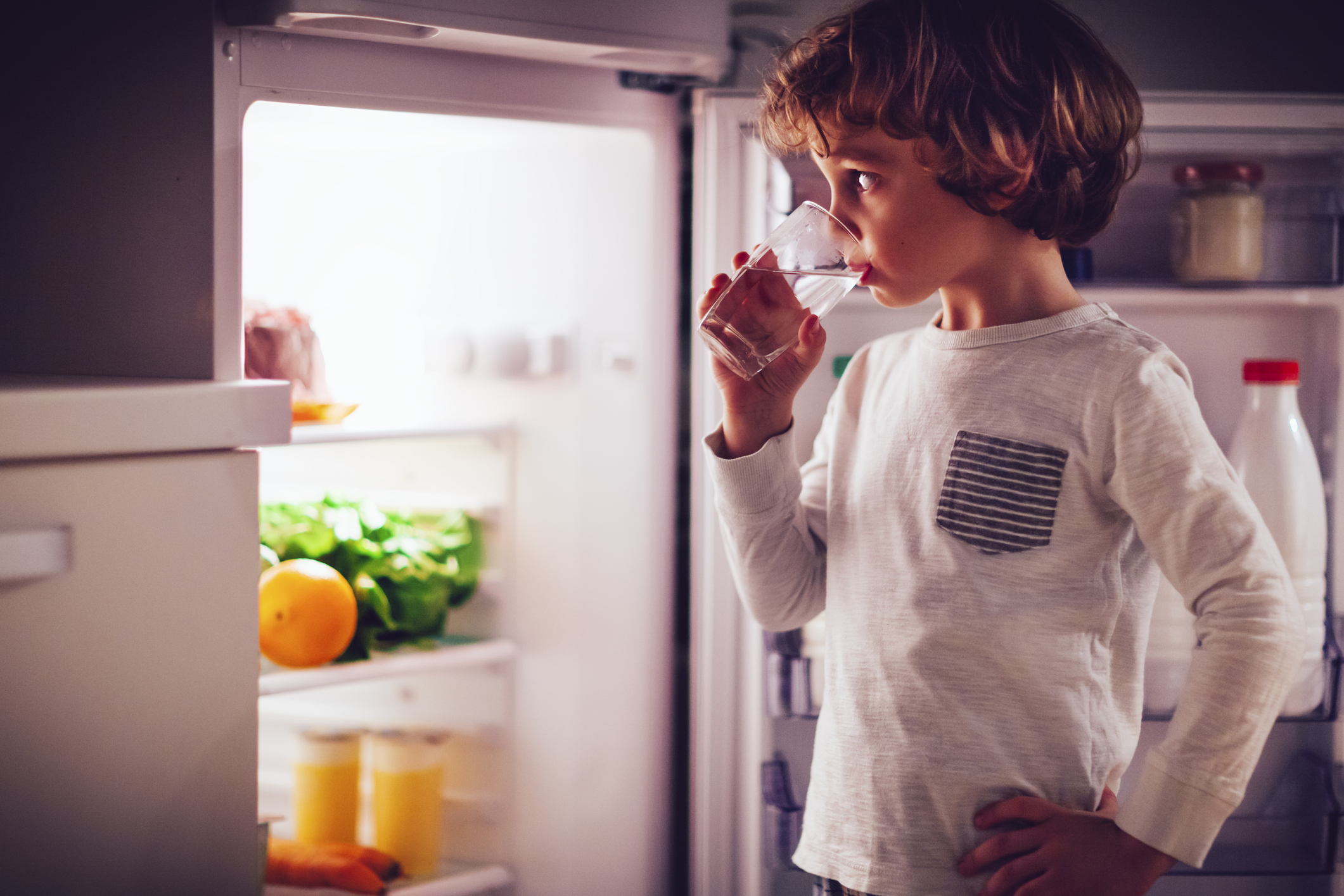 Young boy stands in front of refrigerator drinking glass of water 