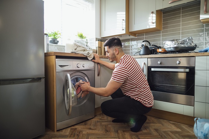 Man crouching down to put clothes into his washing machine in his kitchen.