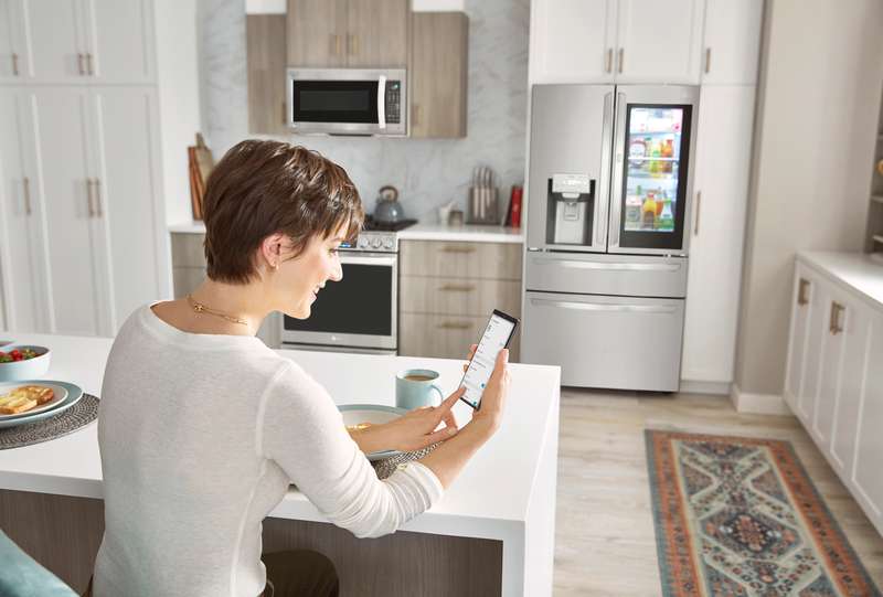 Woman checks her smartphone while eating breakfast in the kitchen with LG smart appliances in the background. v