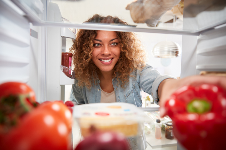 shot taken from inside fridge of young woman smiling while reaching for a red pepper