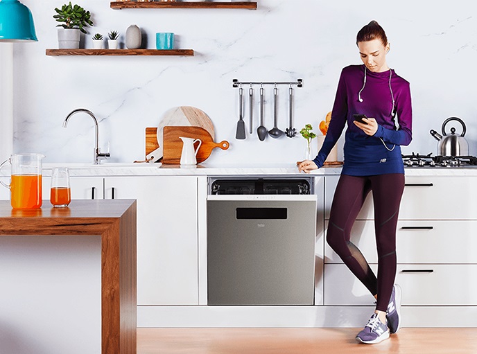 Woman in activewear standing against a modern kitchen counter with headphones on looking at her smartphone while a Beko dishwasher is slightly ajar. 