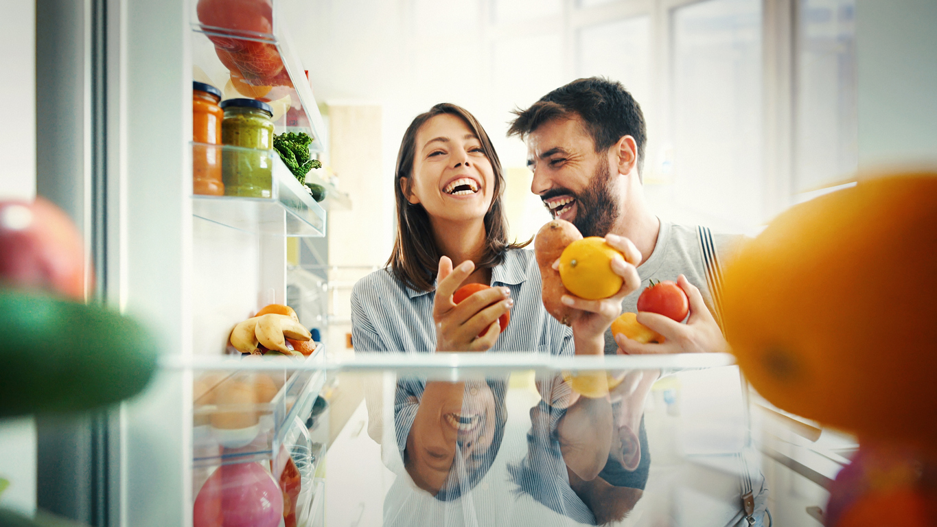Woman and man laughing while they grab produce from inside of a refrigerator