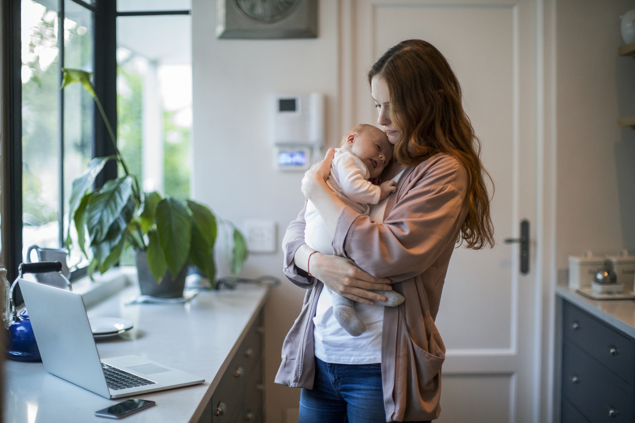 young mother multitasks in kitchen while cradling sleeping infant