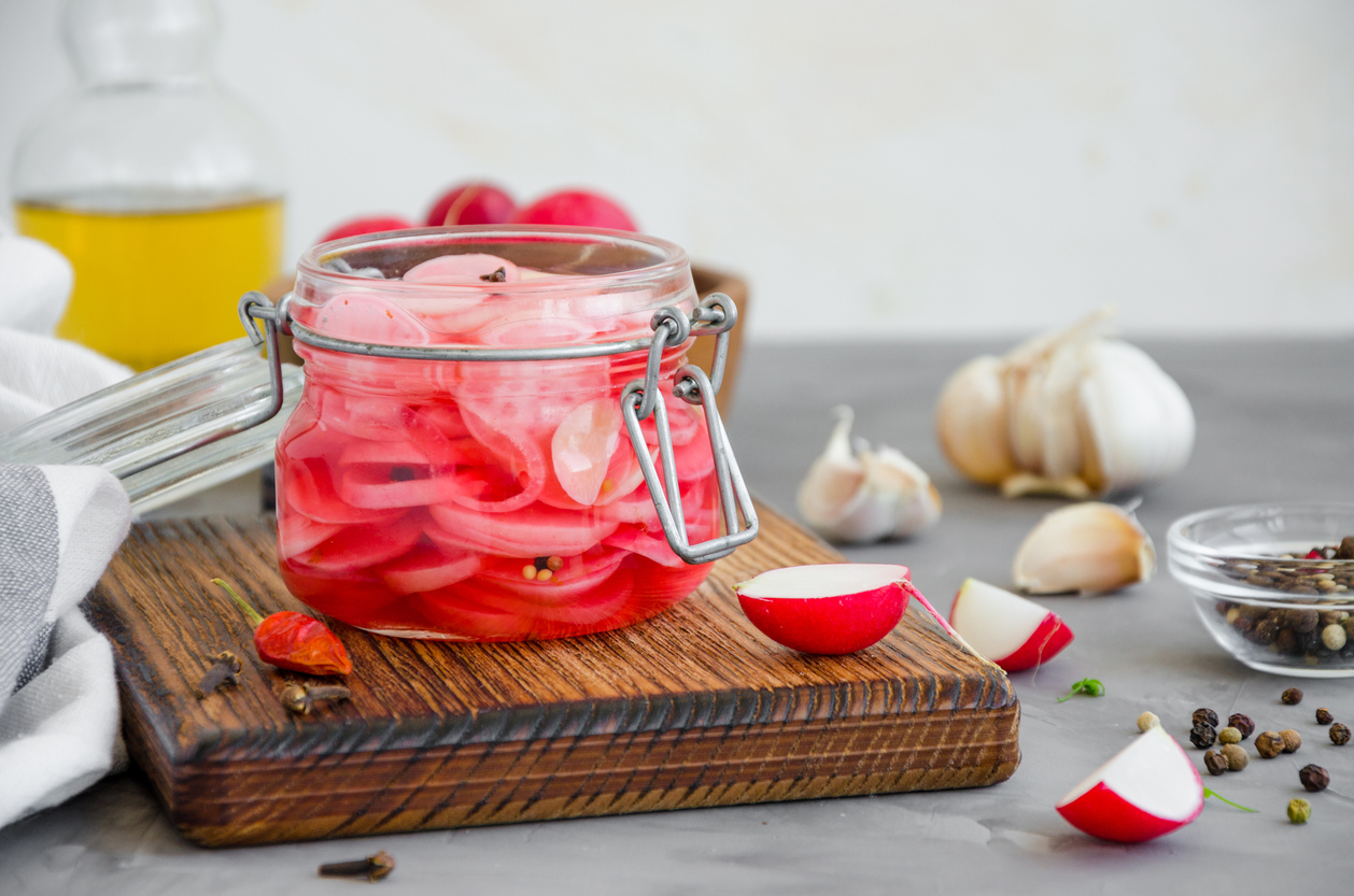 a small glass jar of pickled radishes sits on a cutting board near cloves and garlic 