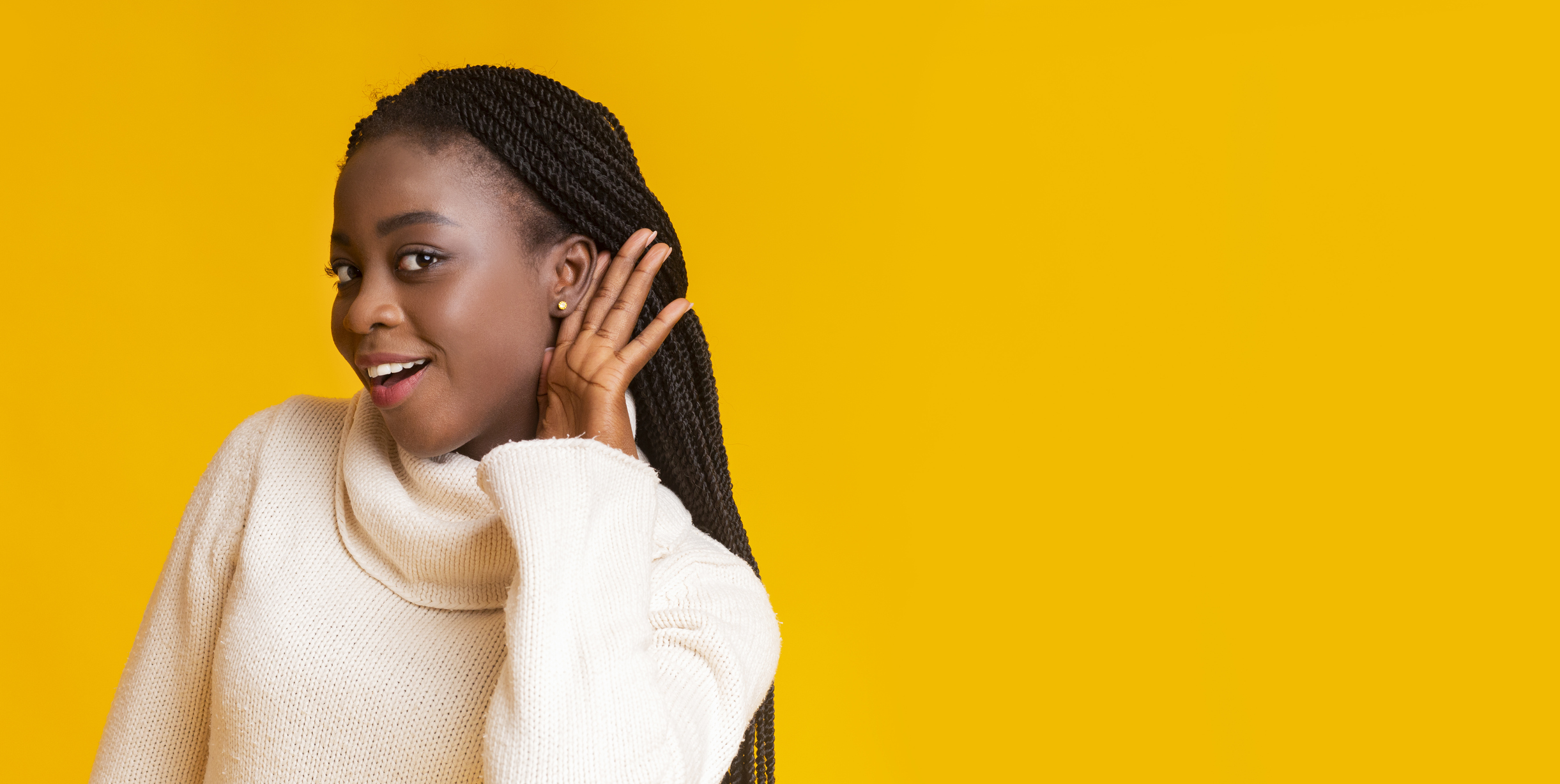 Shot of young African American woman in a turtle neck sweater smiling as she perches her ear to hear a secret against a yellow background 