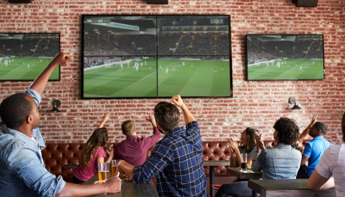 A crowd of people cheering on the soccer game at the pub, with multiple screens