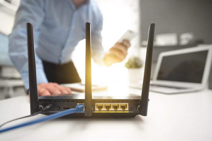 Wireless router with three antennas and cable connected with man using smartphone in background.