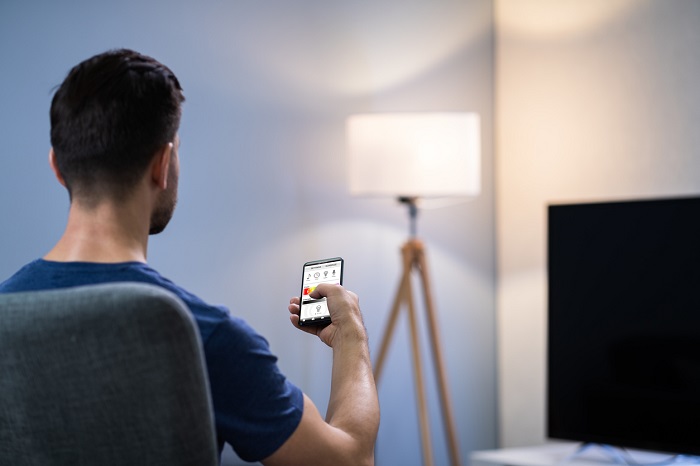 Man sitting in a chair holding up his smart phone as a remote for his lamp in the corner. 