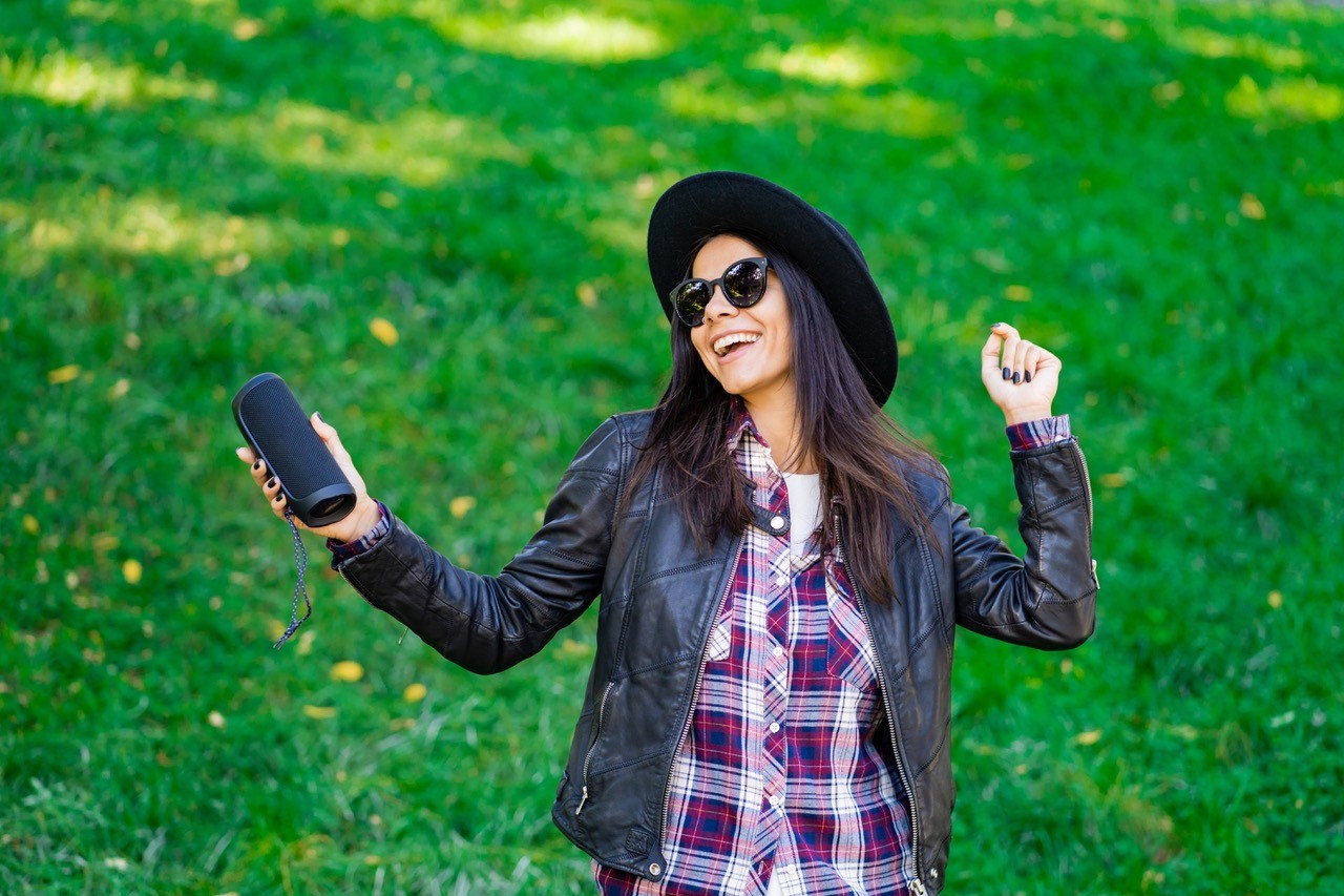 Woman in a hat and jacket joyously listens and grooves to music on her portable speaker outdoors