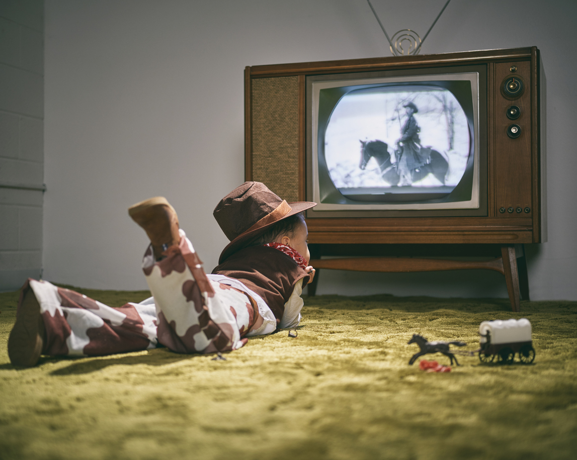 1960s television displaying a cowboy scene on the screen as boy dressed as a cowboy watches