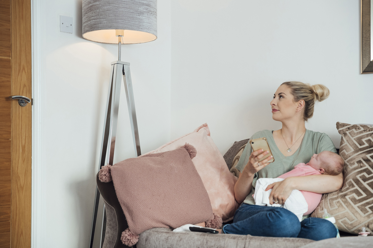 young woman holding her newborn baby in her arms on sofa while turning on the lamp using her phone