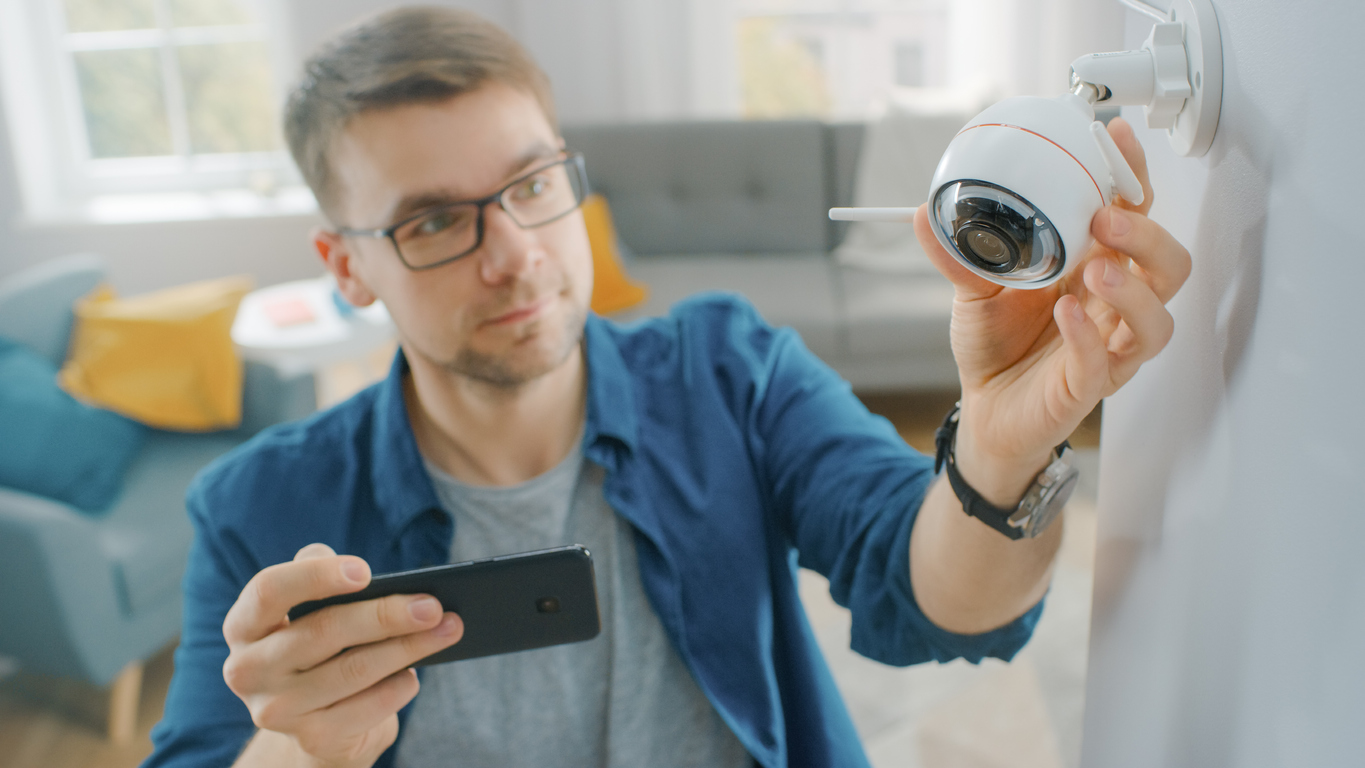 young man adjusting a WiFi surveillance camera at home while checking video feed on smartphone