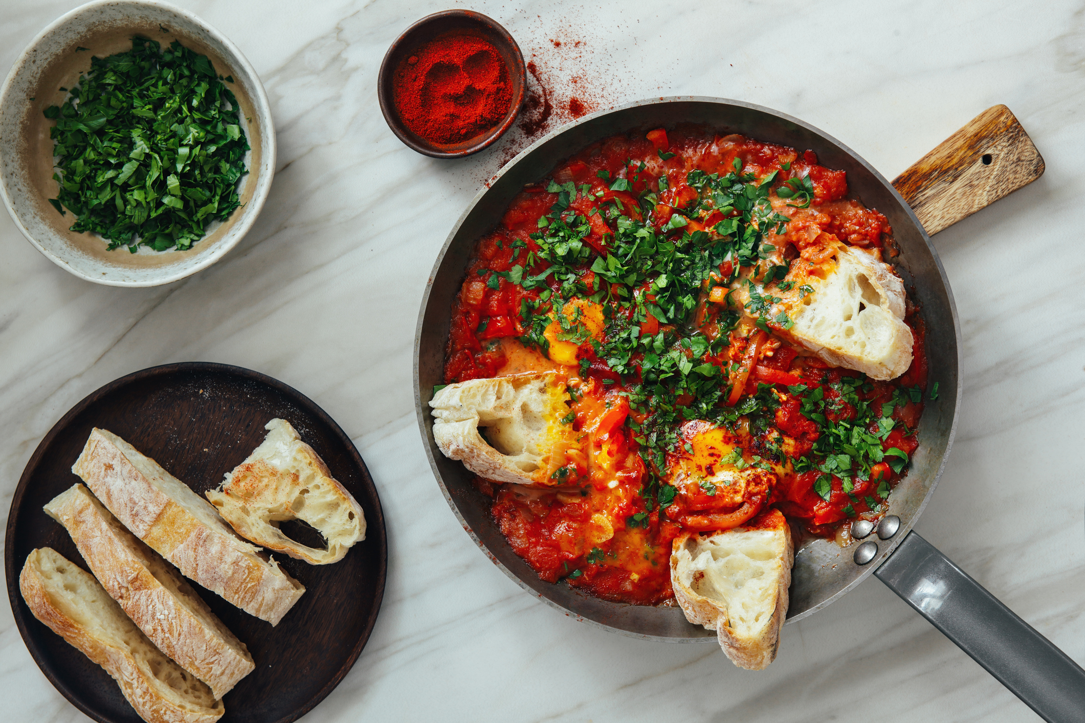 Overhead shot of shakshuka in pan surrounded by herbs, spices, and bread