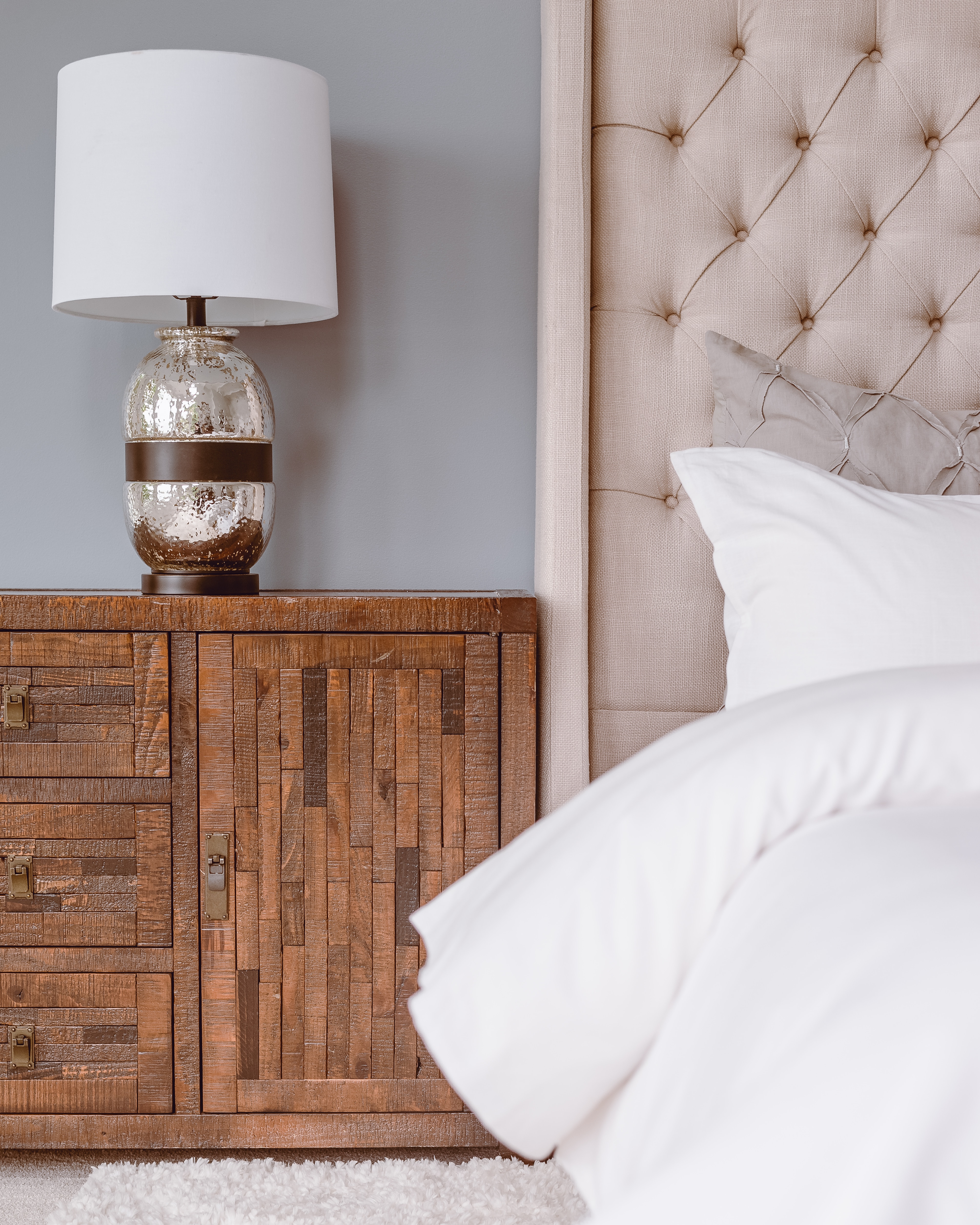 A bed with white linens and an upholstered headboard with a warm wood side table and metallic lamp