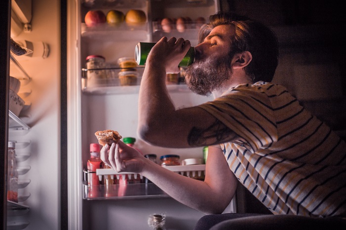 Man with a beard drinking a canned beverage and eating pizza while the refrigerator is open. 