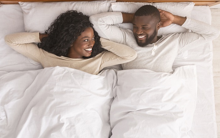 Couple laying next to each other in bed while smiling at each other with their arms behind their heads.