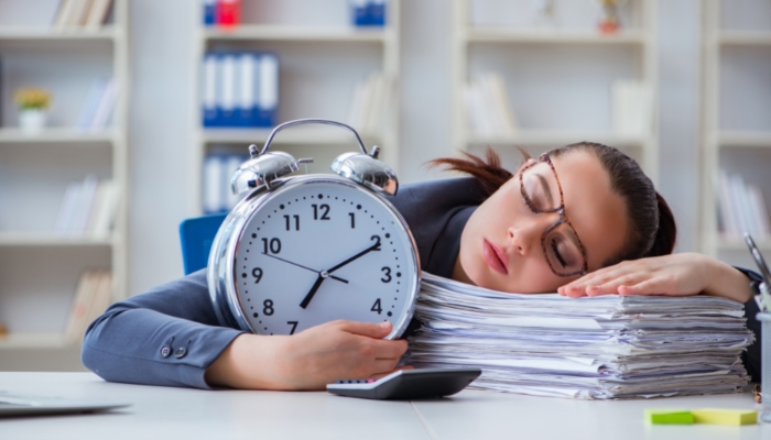 Woman napping at work with alarm clock in hand; indicative of napping duration