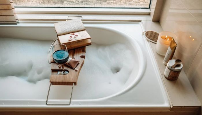 Full white bathtub with bubbles and a bath tray with a book, wine, and lotion
