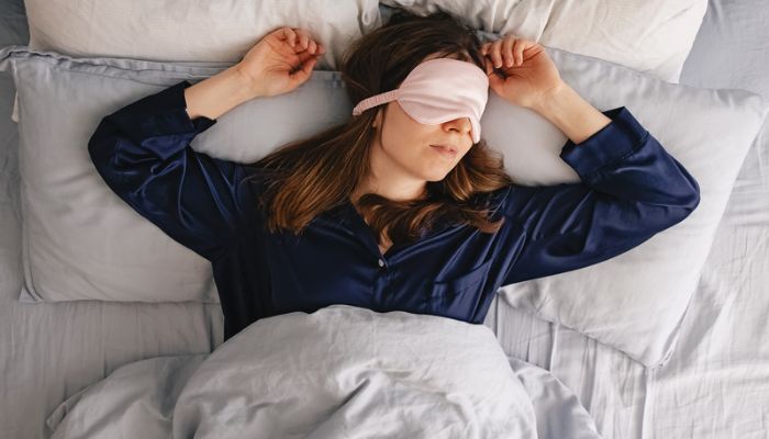Woman in blue pajamas sleeps on her bed with pink eye mask