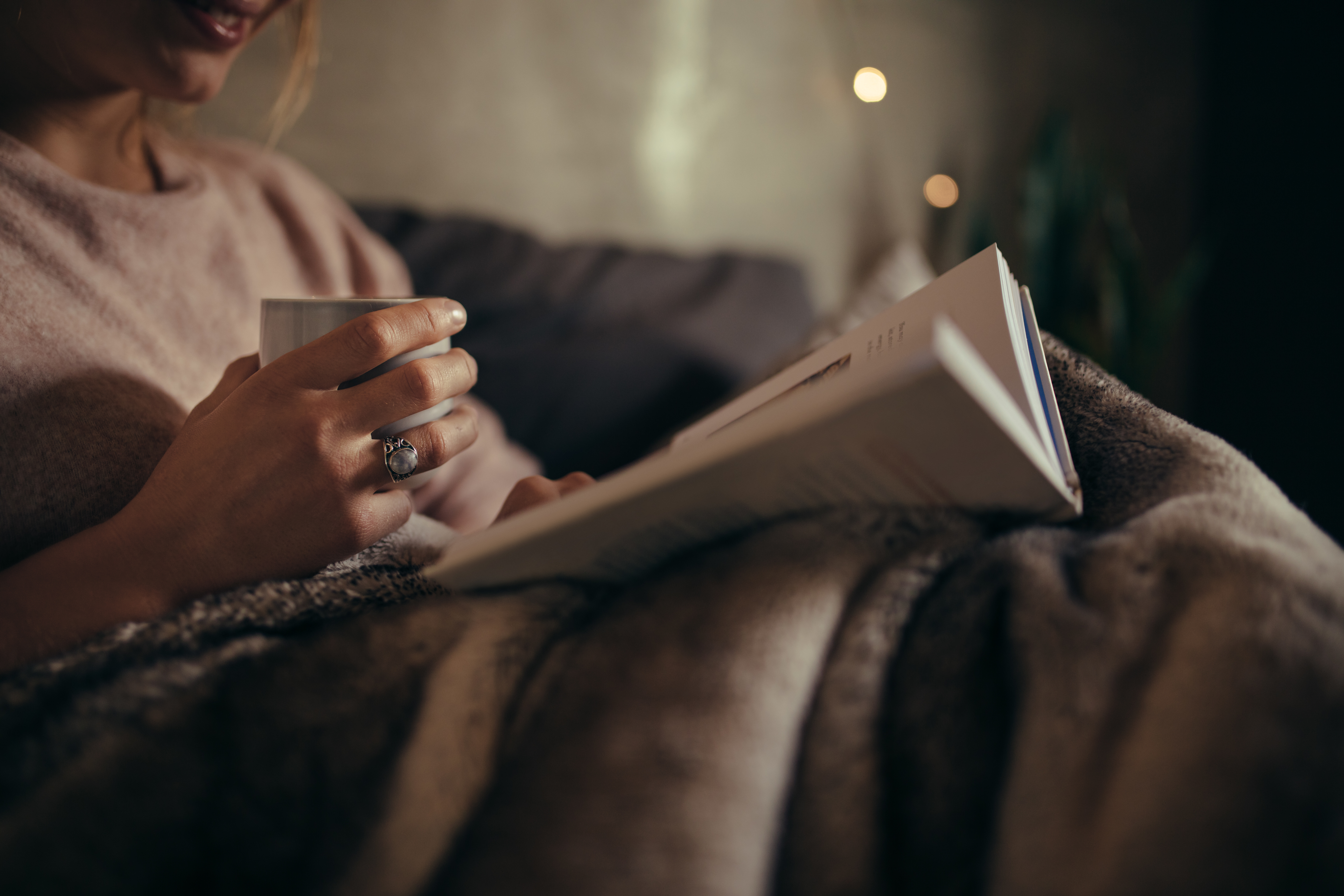 Woman reads a book in bed while wrapped in a blanket and enjoying a hot beverage