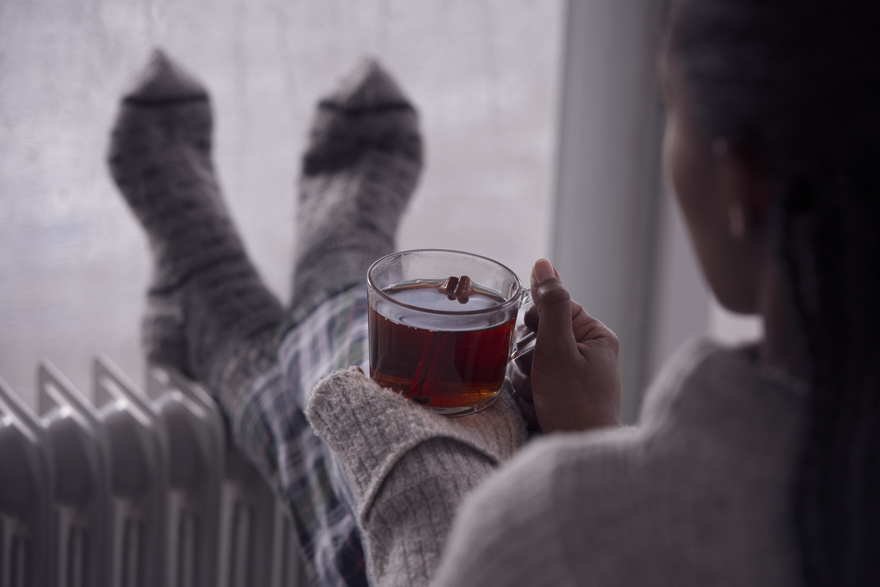 woman drinking hot tea, heating feet on the radiator heater, wearing woolen socks, sitting next to a window