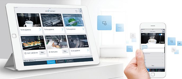 A hand holding a smart phone with the Bosch app open on the screen. A tablet is open on the table next to it, with the Bosch app open as well, connected to various Bosch appliances.
