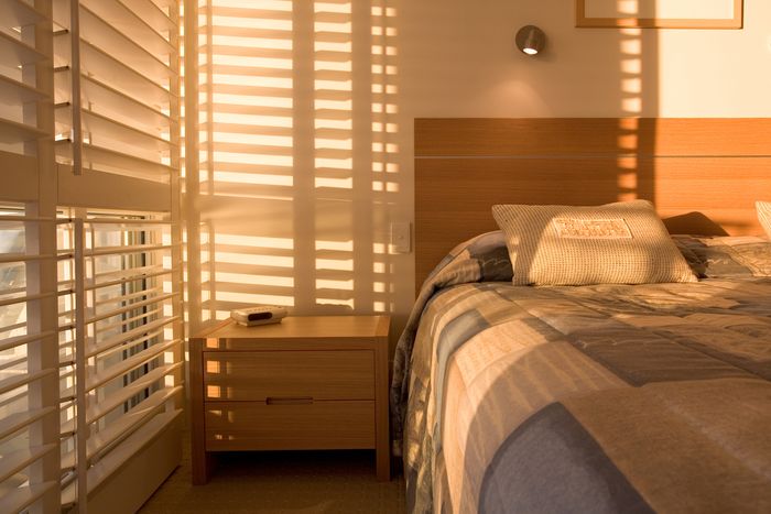 Late afternoon light casts shadows through the slats of a pair of roman shades across a white bedroom, featuring a bed with blue quilted bedspread.