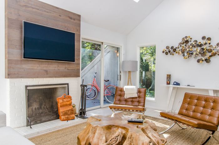 A white living room with dark brown and wood accents. The coffee table is a polished tree stump, and two mid-century modern brown leather chairs stand in front of a flat screen TV.