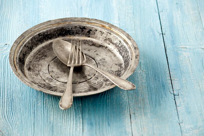 An old, tin dish sits on a blue, wooden table, with tarnished silverware resting inside it.