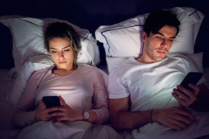A couple laying side by side in bed, both holding phones.