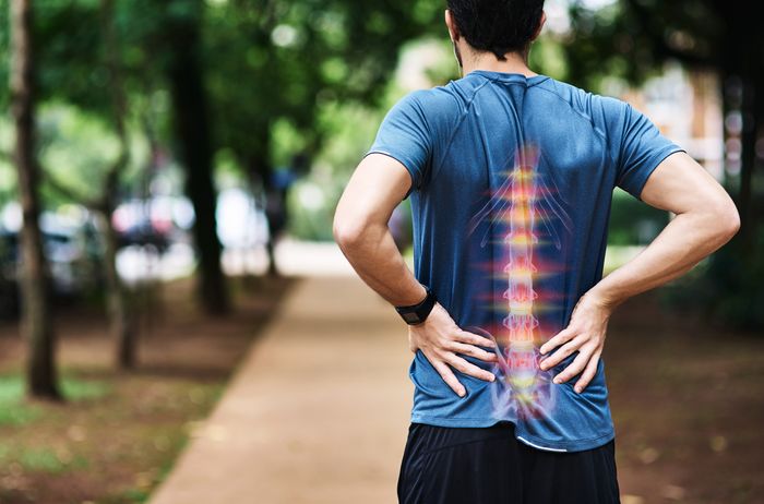 A man stops in the middle of a jog through a park, clutching his back. We can see his spine like an X-ray through the back of his shirt, looking red and inflamed.
