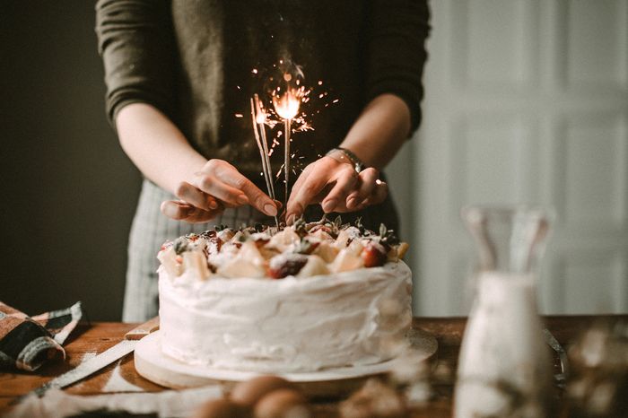 A woman in an earth-toned, rustic kitchen places long, sparkling candles into a vanilla-frosted birthday cake covered in chocolate strawberries.