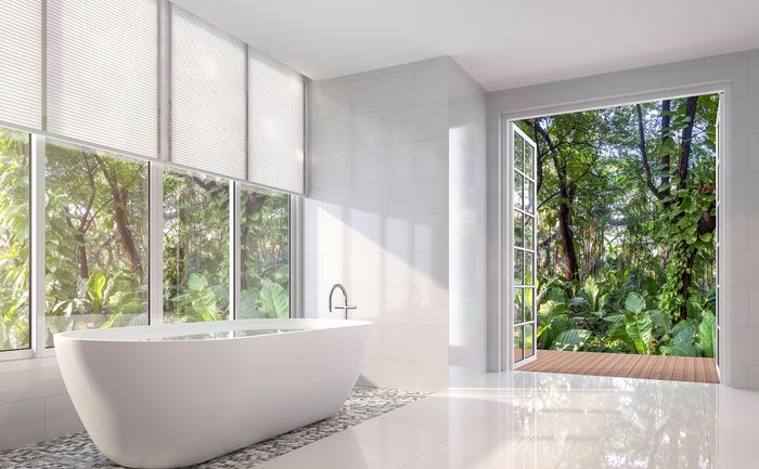 a bright, white bathroom with large, half-shaded windows that show a dense forest outside. Glass doors open up into the woods outside.