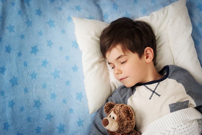 A young boy holds a teddy bear and sleeps on a bed with blue sheets.