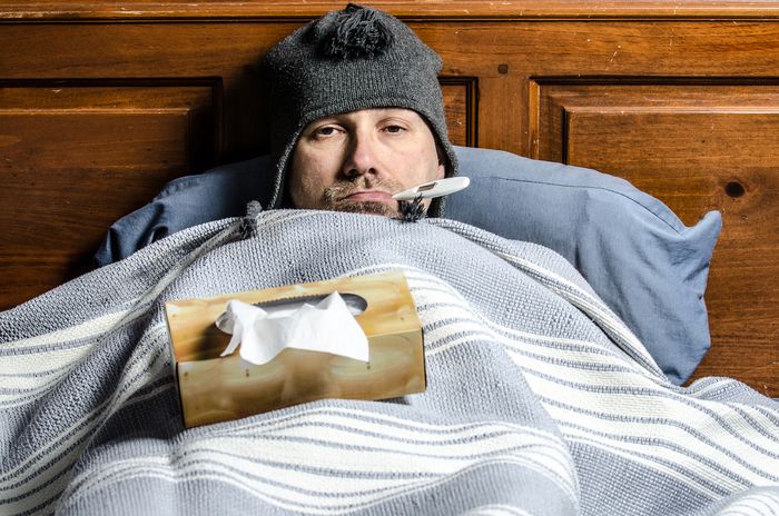 A man in bed wearing a beanie, with a thermometer in his mouth and a box of tissues on top of the blanket.