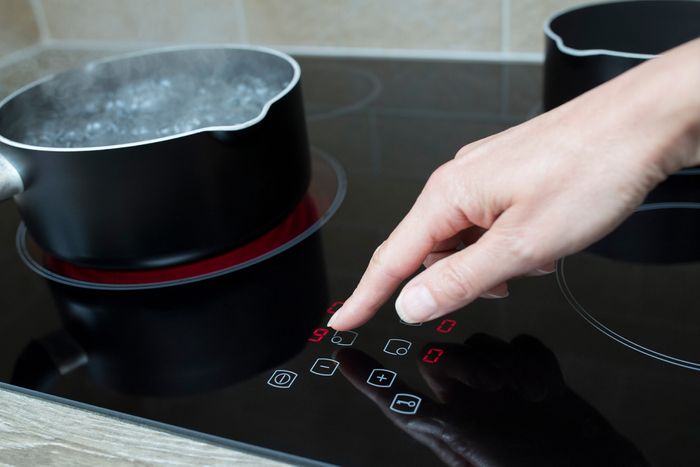 A hand pressing a touchscreen button on a ceramic cooktop, while a pot of water boils on a heating element next to it.